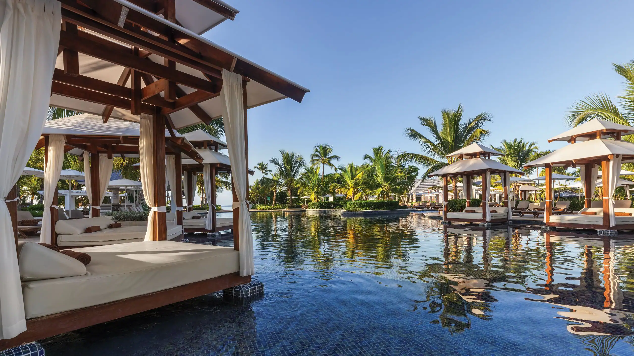 Elegant pool cabanas surrounded by palm trees at Dreams La Romana Resort & Spa overlooking a tranquil Caribbean-style pool.